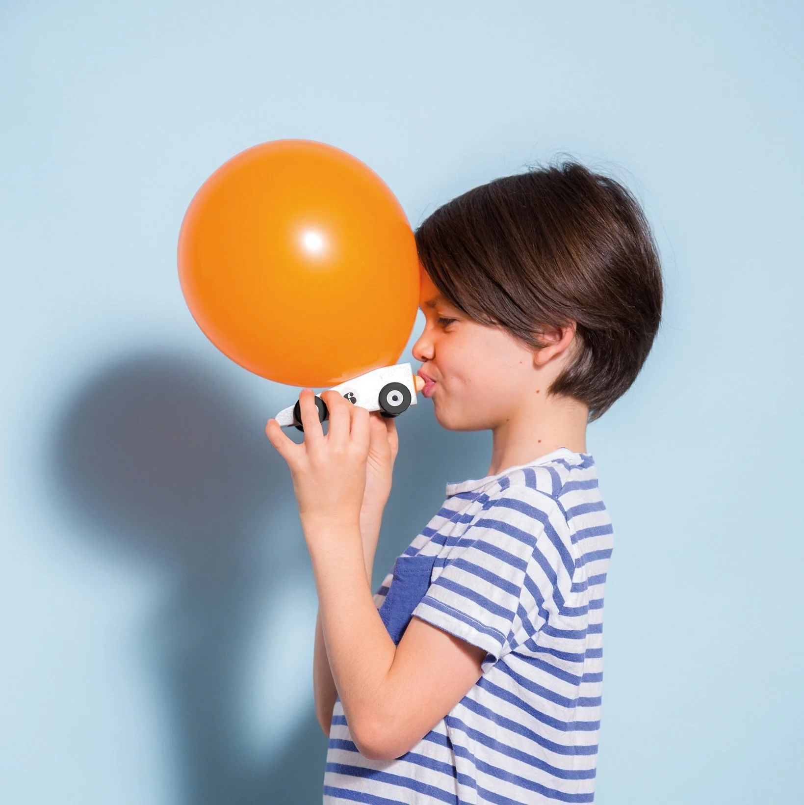 Menino com camisa às riscas segura balão laranja ligado a máquina branca com rodas num fundo azul