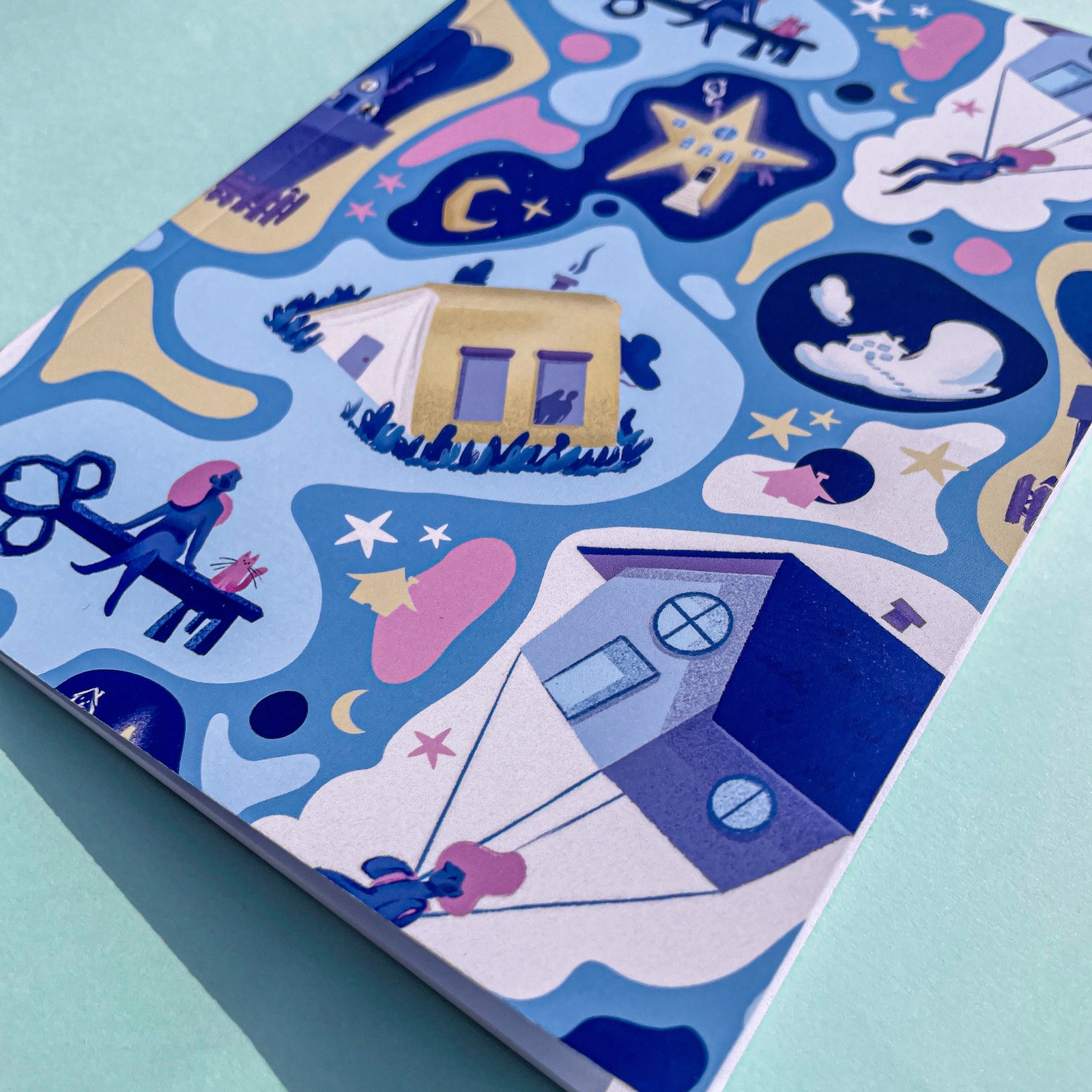 CADERNO A4 "TER UMA CASINHA PARA VIVER" ANDREIA SANTOS Caderno com capa ilustrada com casas, estrelas, guitarra e pessoas em tons azuis e rosa