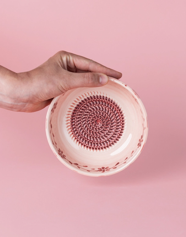 TAÇA RALADOR MÉDIA "SAKURA" ALFAR TIERRA COCIDA Tigela cerâmica branca e vermelha com padrão em espiral e flores, mão segurando, fundo rosa