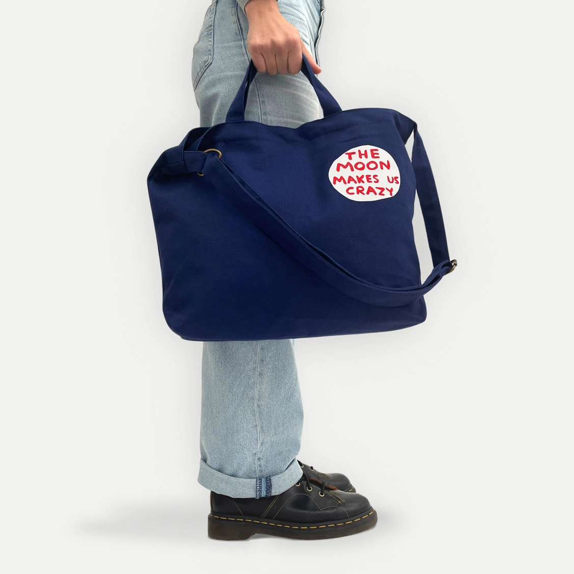 BOLSA "THE MOON MAKES US CRAZY" - DAVID SHRIGLEY Mala azul escura com texto THE MOON MAKES US CRAZY sendo segurada por pessoa com jeans claros e sapatos pretos.