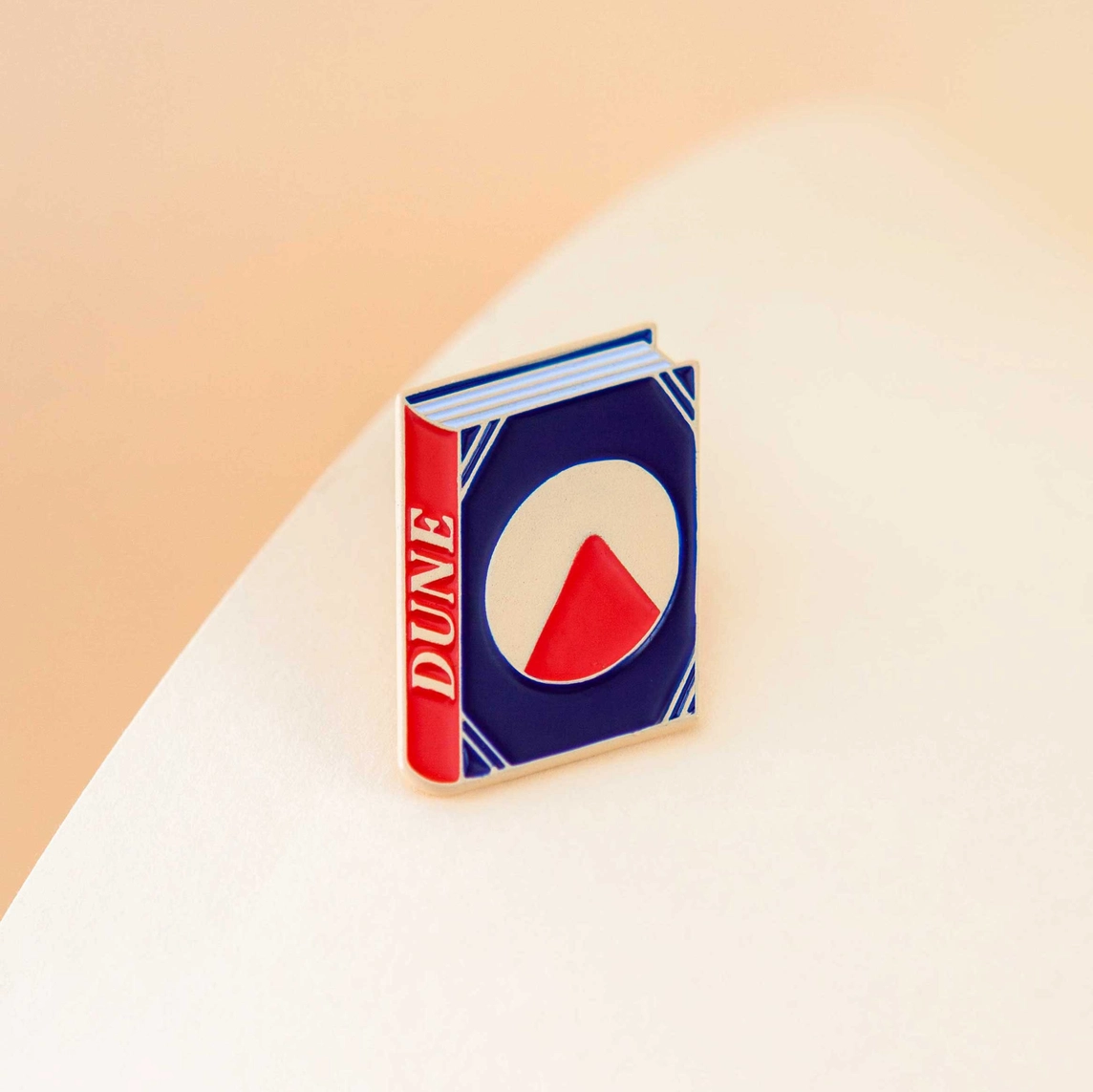 PIN "DUNE BY FRANK HERBERT" JUDY KAUFMANN Livro DUNE com capa azul, vermelho e branco sobre superfície clara.