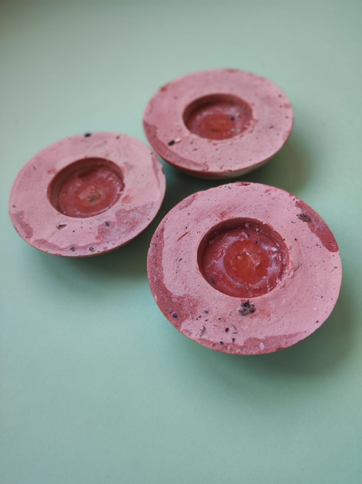 CONJUNTO DE 3 CASTIÇAIS RASOS PARA VELAS "TABLEWARE FOR ISOLATION" MARQUES MARQUES Três velas rosa com manchas castanhas em fundo verde claro.