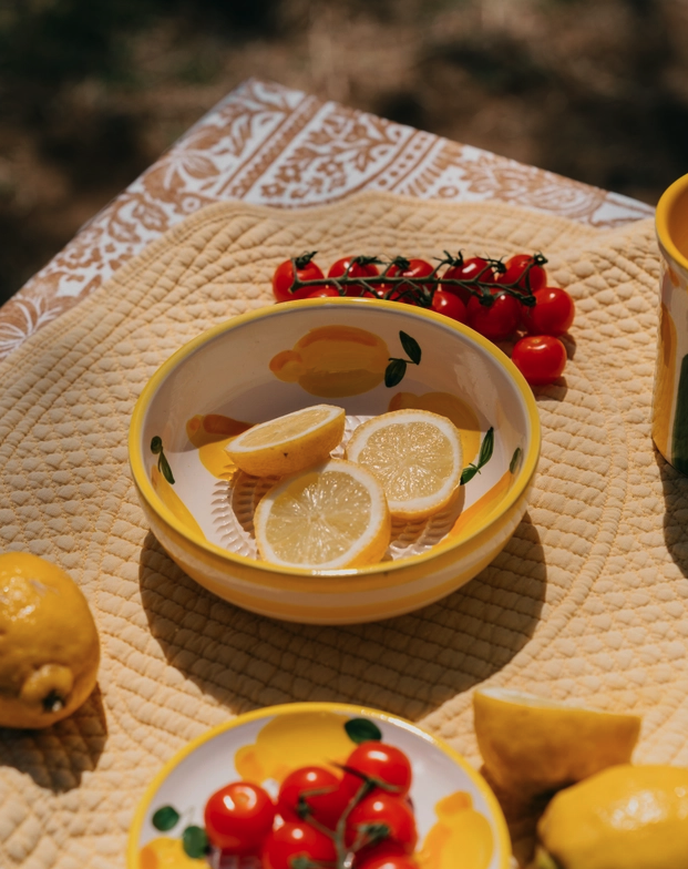 TAÇA RALADOR MÉDIA "LIMONE" ALFAR TIERRA COCIDA Taça e prato de cerâmica com limões e tomates cherry sobre tecido acolchoado e toalha estampada