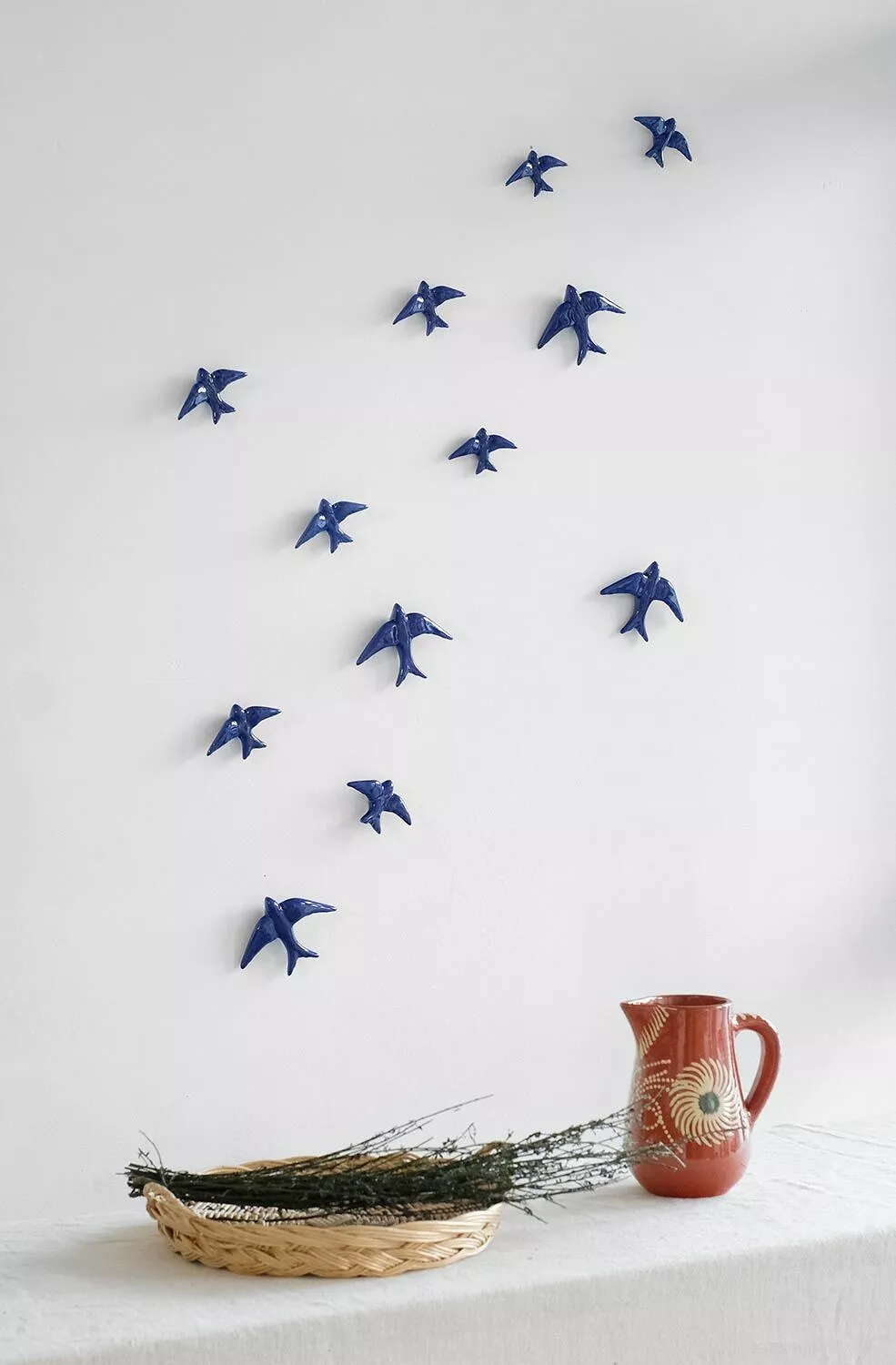 GLAZED COBALT BLUE SWALLOW CASA ATLANTICA Conjunto de pássaros decorativos azuis na parede com jarra cerâmica vermelha e tabuleiro de palha com ramos secos numa superfície branca
