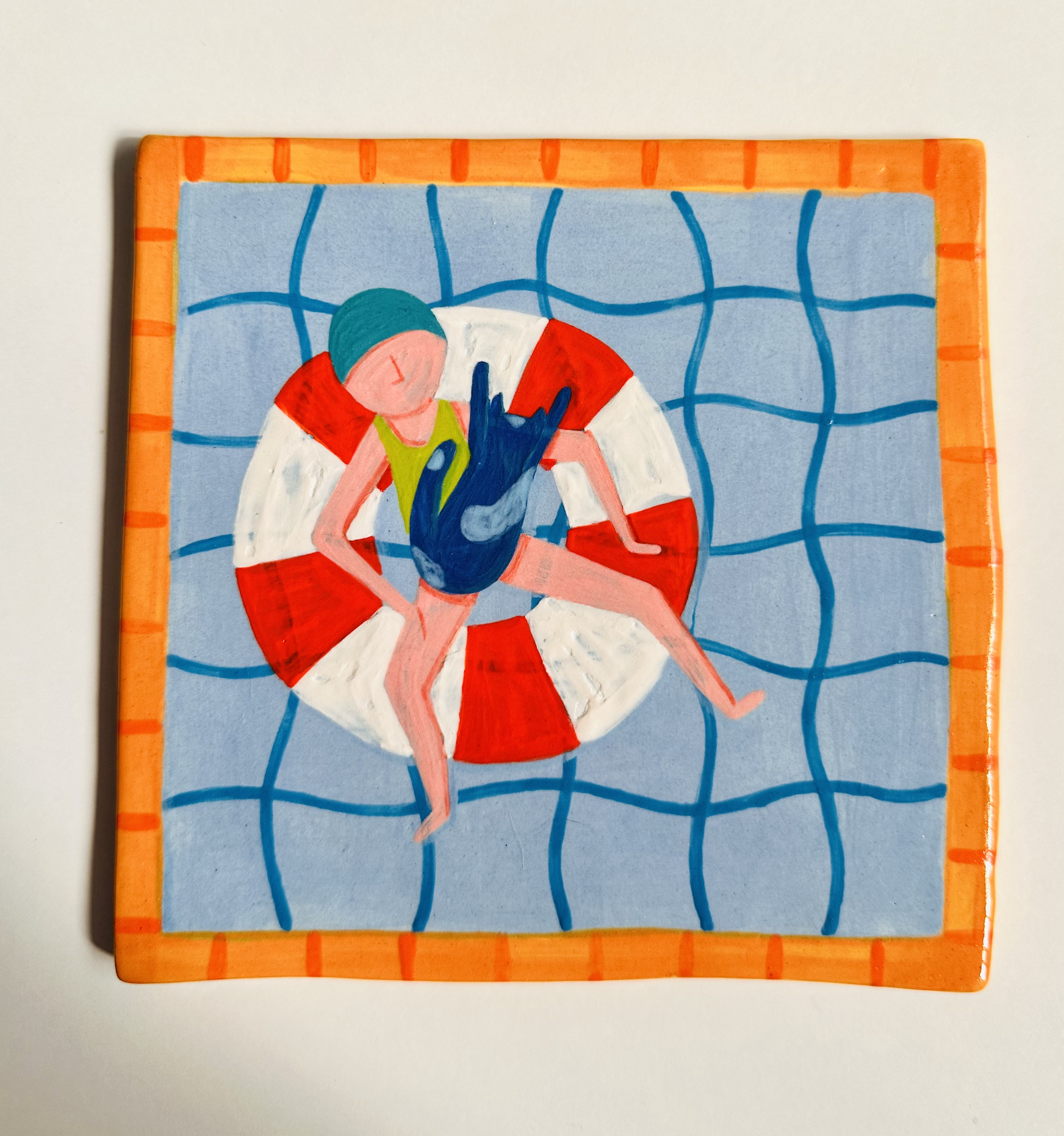 AZULEJO PISCINA BOIA GATO KIKA 14X14CM Azulejo com figura de pessoa com roupa de banho azul num salva-vidas vermelho e branco, fundo azul com padrão em grelha e borda laranja.