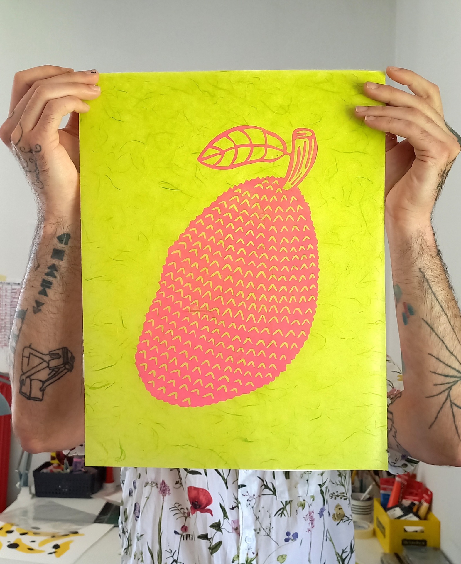 LINOGRAVURA "JACA" JEFFERSON RIB Cartaz de papel verde fluorescente com ilustração cor-de-rosa de uma manga, segurado por pessoa com tatuagens e camisa floral.