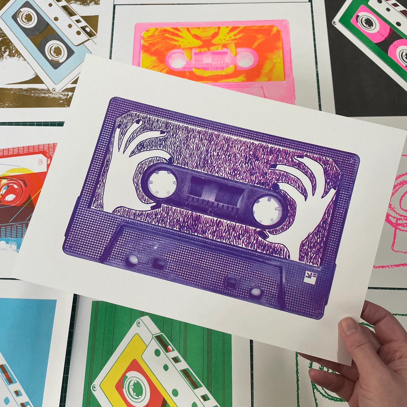 RISO MIXED TAPES - HANDS ON TAPE - A4 VRIJFORMAAT Impressão artística de cassete roxa com desenho de mãos em fundo de outras impressões de cassetes coloridas