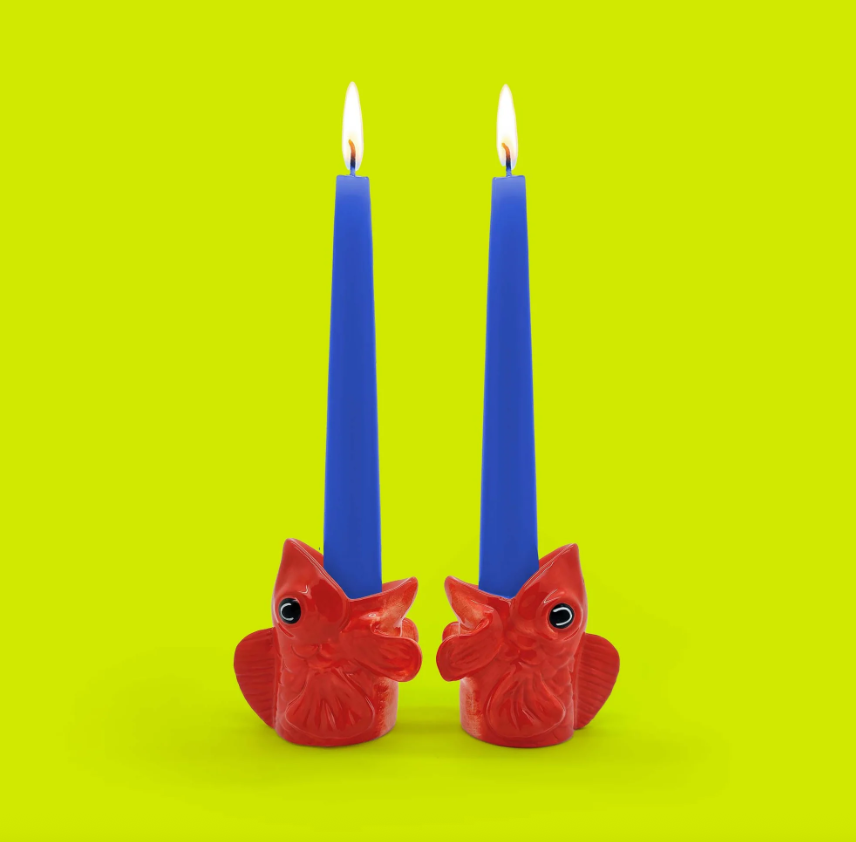 CANDLEHOLDER SET "FISHES FOR DISHES" DONKEY Dois castiçais vermelhos em forma de cabeça de peixe com velas azuis acesas em fundo amarelo.
