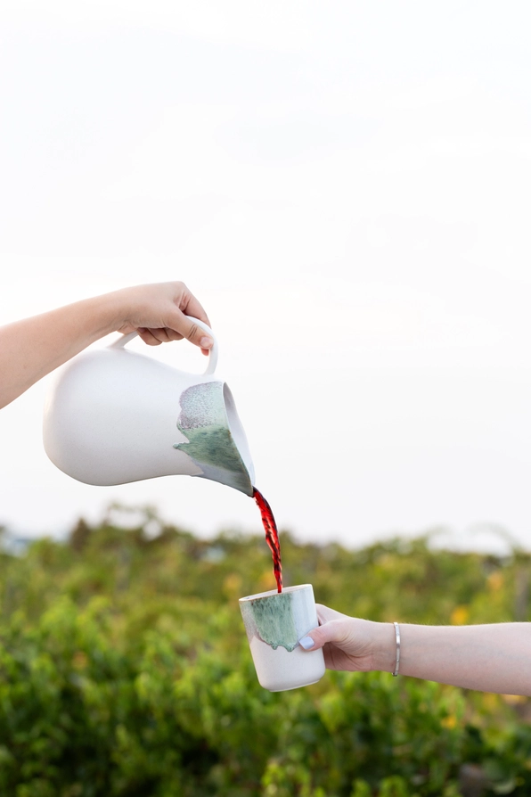 JARRO DOURO EGG BACK HOME Jarro branco a servir vinho tinto em caneca branca com detalhes verdes ao ar livre