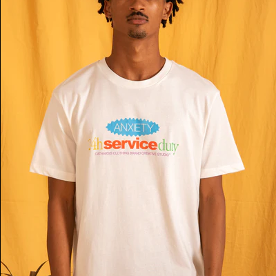 T-shirt branca com texto colorido ANXIETY 24h service duty, homem jovem, fundo amarelo e tangerinas na mesa