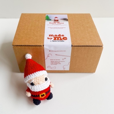 Boneco de croché de Pai Natal junto a uma caixa de cartão com etiqueta branca estampada