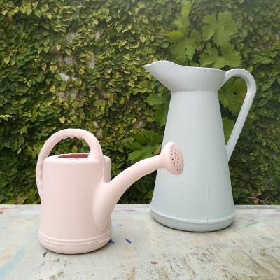 Regador rosa e jarro cinza em superfície clara com fundo de vegetação