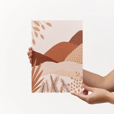 Quadro decorativo com ilustração de paisagem em tons terra sendo segurado por uma pessoa