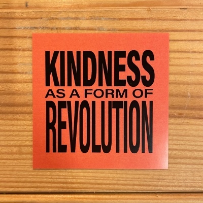 Autocolante quadrado laranja com texto a preto 'KINDNESS AS A FORM OF REVOLUTION' sobre madeira clara