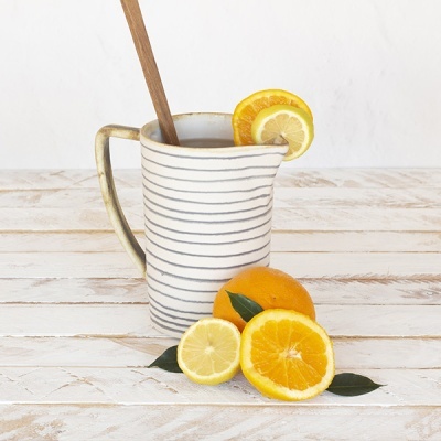 Jarra branca com riscas cinzentas, colher de pau e frutas cítricas em mesa de madeira