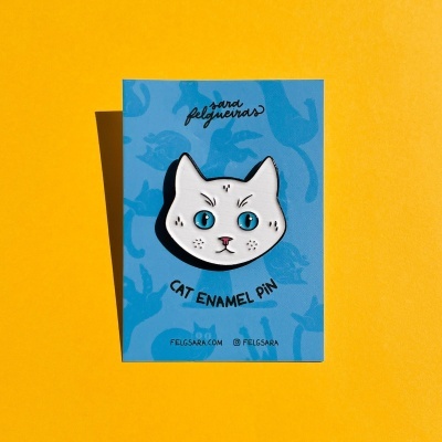 Pin esmaltado da cabeça de um gato numa cartolina azul com texto, fundo amarelo