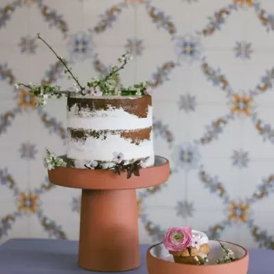 Bolo decorado com flores num suporte cerâmico laranja sobre mesa azul e azulejos decorativos ao fundo