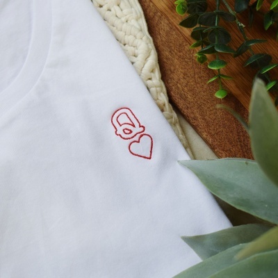 Camisola branca com bordado vermelho de cadeado com coração, sobre madeira e tapete creme com plantas verdes