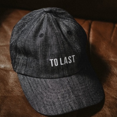 Boné cinza escuro com texto branco 'TO LAST' sobre superfície de couro marrom