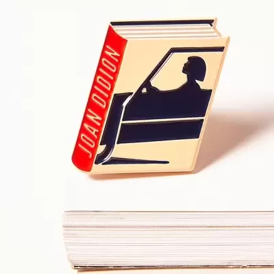 Pin metálico em forma de livro com desenho de pessoa a conduzir e texto JOAN DIDION