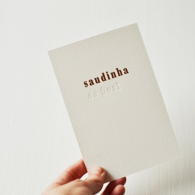 Cartão branco texturizado com textos 'saudinha' e 'de toal', segurado por mão.