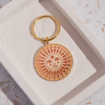 Chaveiro metálico dourado com rosto de sol estilizado e prato branco de cerâmica