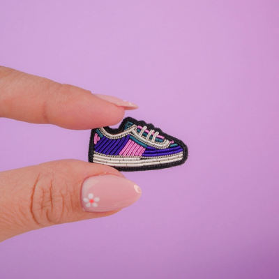 Aplicação bordada de sapatilha colorida segurada por dedos femininos com fundo lilás.