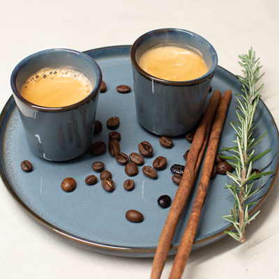Duas chávenas pequenas de café expresso num prato azul com grãos de café, canela e alecrim