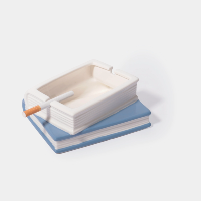 Cinzeiro cerâmico branco em forma de livro sobre livro azul com cigarro