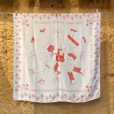 Pano branco quadrado com bordado vermelho de figura humana e texto pendurado numa corda