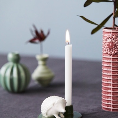 Castiçal verde com vela branca e ouriço branco, vasos decorativos ao fundo.