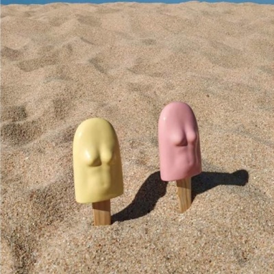 Dois gelados em paus; amarelo e rosa; forma de corpo feminino; na areia da praia; céu azul claro