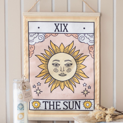 Quadro têxtil com ilustração do Sol com rosto e texto 'THE SUN' pendurado numa parede de madeira.