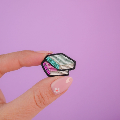 Broche de livros bordados segurado entre dedos com unha decorada em fundo roxo
