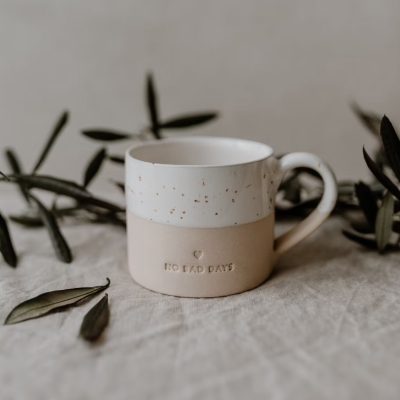Caneca cerâmica branca e bege com texto NO BAD DAYS sobre tecido cinza e folhas verdes