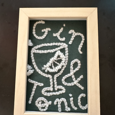 Quadro com moldura de madeira e bordado branco em fundo verde com frase 'Gin & Tonic' e desenho de copo.