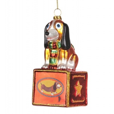 Enfeite de Natal em vidro com cão sentado sobre cubo laranja com estrelas douradas.