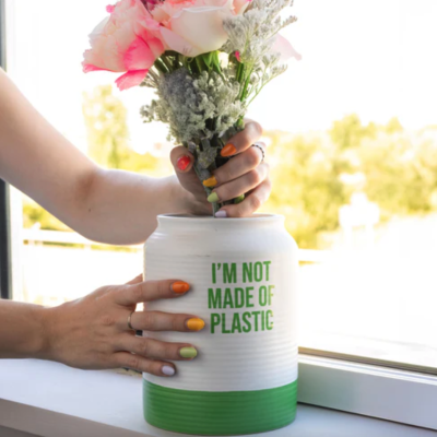 Vaso cerâmico branco e verde com flores e texto em inglês, mãos segurando o vaso numa janela