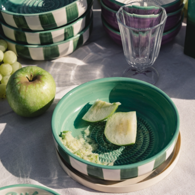 Tigelas de cerâmica decoradas com padrões verdes, brancos e roxos e uma taça de vidro transparente numa mesa clara