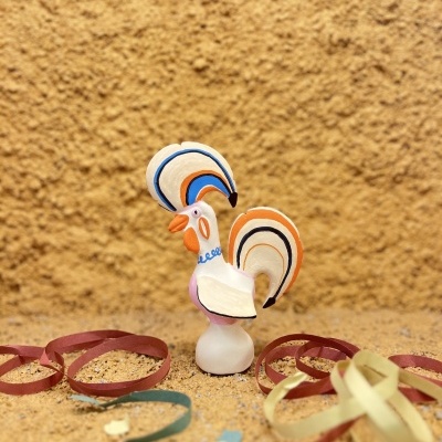 Figurinha cerâmica pintada de galo com detalhes coloridos e fitas coloridas ao redor sobre superfície castanha texturada.