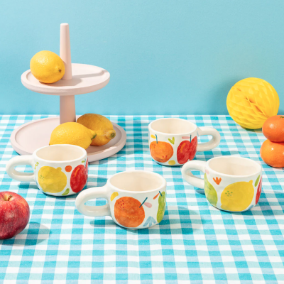 Quatro chávenas de cerâmica com padrão de frutas coloridas sobre toalha xadrez azul e branca com frutas e suporte de dois andares