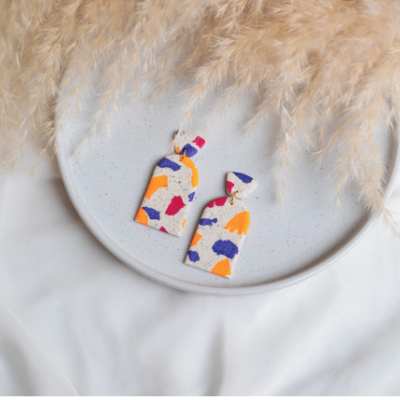 Brincos coloridos em padrão geométrico sobre prato branco com penas secas ao lado