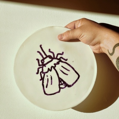 Prato branco com desenho bordado roxo segurado por mão com tatuagem.