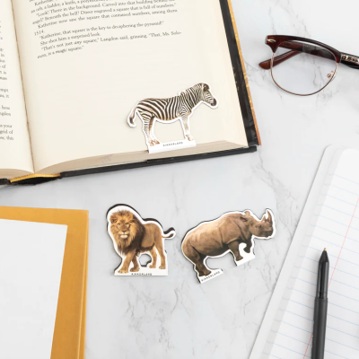 Marcadores de página com forma de zebra, leão e rinoceronte sobre mesa com livro aberto, caderno, caneta e óculos