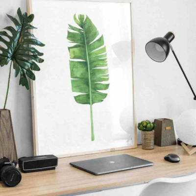 Mesa de trabalho com laptop, candeeiro, planta e quadro com ilustração de folha verde