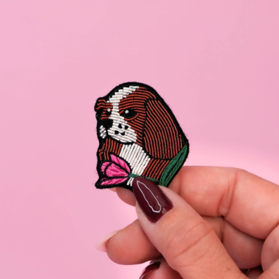 Emblema bordado de cão castanho e branco segurado pela mão com unhas cor vinho, fundo rosa