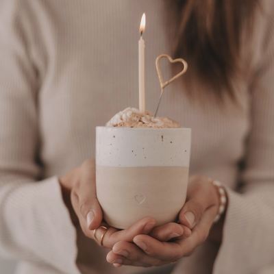 Vaso de cerâmica com vela acesa e decoração em arame em forma de coração, segurado por mãos femininas.
