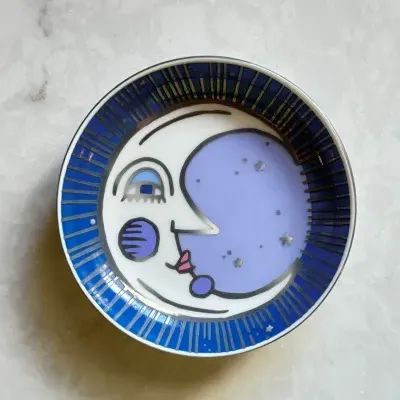 Prato redondo decorativo com desenho de lua azul e branca com detalhes coloridos em superfície clara.