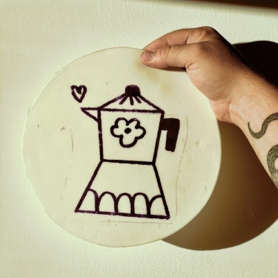 Disco redondo branco com desenho preto de cafeteira, mão tatuada segura