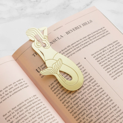 Marcador de livros dourado em formato de sereia colocado em livro aberto sobre superfície de mármore
